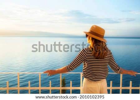 Shot of woman wearing straw hat and sunglasses while standing on balcony and looking at sea view. Daydreaming.