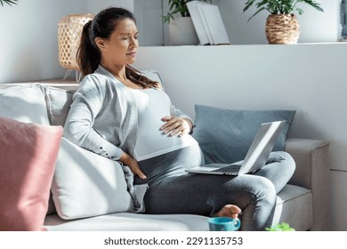 Shot of worried pregnant woman with a stomachache working with laptop sitting on sofa in the living room at home - Powered by Shutterstock