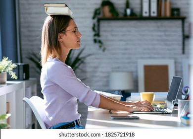 Shot Of Young Attractive Woman At The Desk With Books On Her Head While Working With Computer At Home.