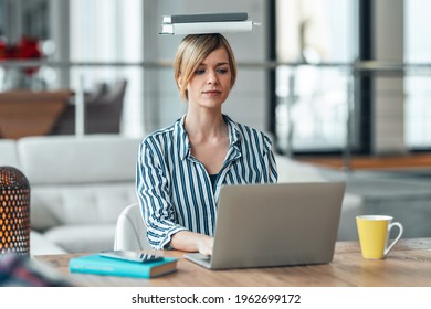 Shot Of Young Attractive Woman At The Desk With Books On Her Head While Working With Computer In Living Room At Home.