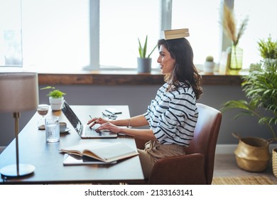 Shot of young attractive woman at the desk with books on her head while working with computer at home. Portrait of attractive woman at desk, books on her head