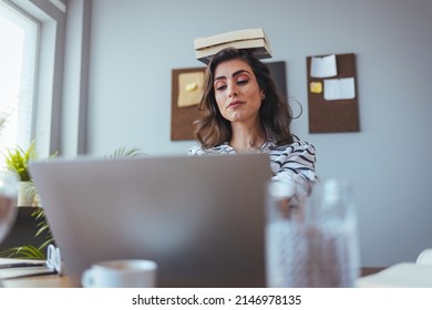Shot of young attractive woman at the desk with books on her head while working with computer at home. Portrait of attractive woman at desk, books on her head