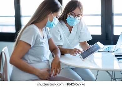 Shot of young beautiful gynecologist wearing a hygienic face mask while showing to pregnant woman ultrasound scan baby with digital tablet in medical consultation.