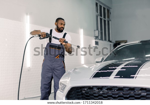 Shot of young black man in working\
clothes, car wash employee, washing the car hood under high\
pressure water gun at professional auto washing\
service.