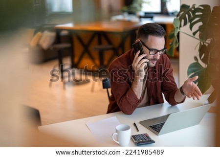 Shot of a young businessman talking, having a discussion on a cellphone, explaining the current situation while working, looking on a laptop in a home office.