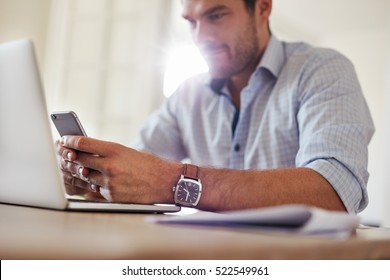 Shot Of Young Businessman Using Mobile Phone At Home. Man Sitting At Table With Laptop And Reading Text Message On His Smart Phone.