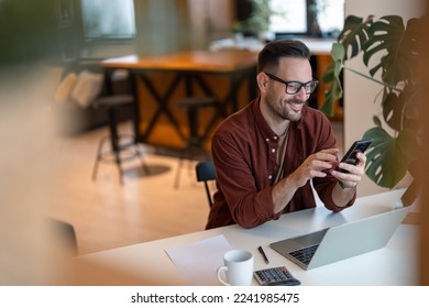 Shot of a young businessman using a smart phone in a home office. Smiling, touching the screen, browsing the internet. - Shutterstock ID 2241985475