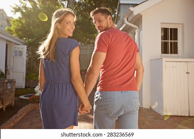 Shot Of Young Couple In Backyard On A Bright Sunny Day. They Are Walking To Their House Hand In Hand, Looking Over Their Shoulders At The Camera.