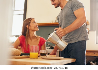 Shot Of Young Couple Having Breakfast In Kitchen. Young Man Standing And Serving Coffee With Woman Sitting By Breakfast Table At Home In Domestic Kitchen.