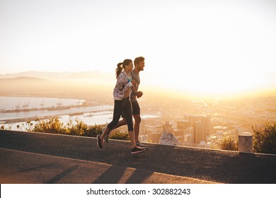 Shot Of Young Couple Running On Hillside Road Outside The City.  Young Man And Woman Jogging In Morning With Bright Sunlight.