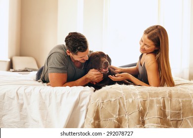 Shot Of A Young Couple With Their Dog On The Bed In Morning. Young Man And Woman Spending Time With Their Pet In Bedroom.