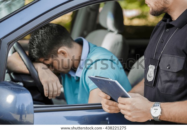 Shot of a young driver\
keeping his head on a steering wheel and a policeman standing next\
to the car