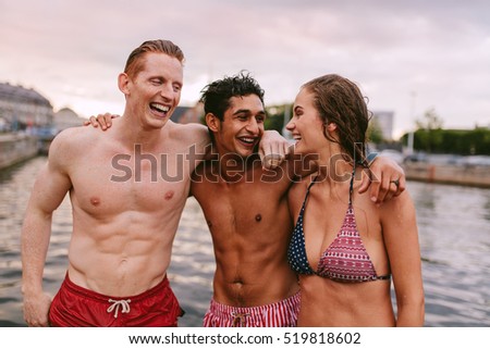 Shot of young friends in swimwear standing together by the lake and laughing. Men and woman  enjoying a day by the lake.