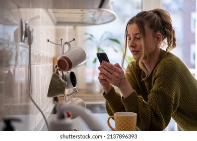 Shot of a young woman drinking tea and using smartphone in her kitchen while getting ready to go to work. Watching videos or messaging with friends while enjoying morning at home. - Shutterstock ID 2137896393