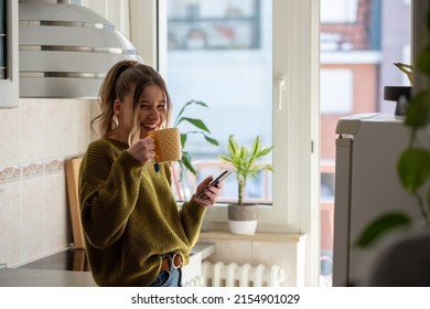Shot of a young woman drinking tea and using smartphone in her kitchen while getting ready to go to work. Watching videos or messaging with friends while enjoying morning at home. - Powered by Shutterstock