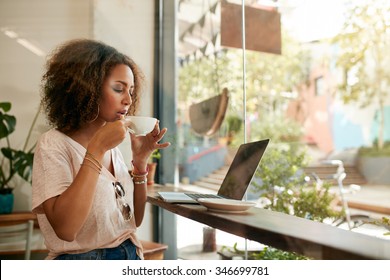 Shot of young woman sitting in a coffee shop with laptop and drinking coffee. African american girl having hot and fresh coffee at cafe.