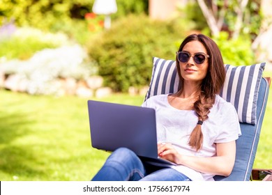 Shot Of Young Woman Sitting On Sunbed In The Garden And Using Her Laptop.