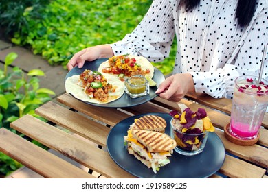 Show food in hands, woman's hand picking plate of Mexican tacos food with half of cut sandwich in black dish on wooden table in the garden, iced sweet beverage, brunch or breakfast  - Powered by Shutterstock