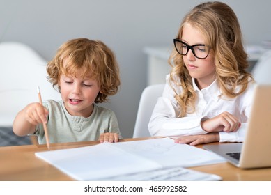 Show Me. Prudent Little Girl With Glasses Looking At Little Adorable Boy Holding A Pencil And Drawing While Sitting At The Table