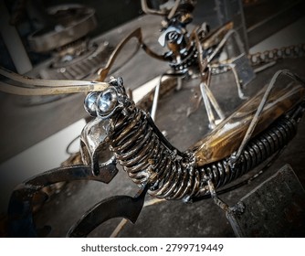showcases a dramatic, close-up view of an intricate insect sculpture crafted from welded scrap metal parts like coiled springs and polished bearings, while a second mechanical creature rests softly blurred in the background.
