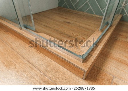 Shower stall threshold. The floor is made of wood-look tile and the shower is enclosed in glass. The edge of the shower stall has a thin, silver trim. Glass door hinges.