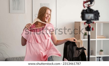 Showing clothes. Filming video. Fashion advice. Cheerful playful young woman trying pink dress on hanger recording vlog on camera on tripod in white room.