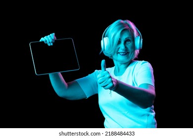 Showing Device Screen. Emotional Senior Woman In White T-shirt Using Digital Tablet Isolated On Dark Background In Neon Light. Concept Of Beauty, Age, Emotions, Modern Gadgets