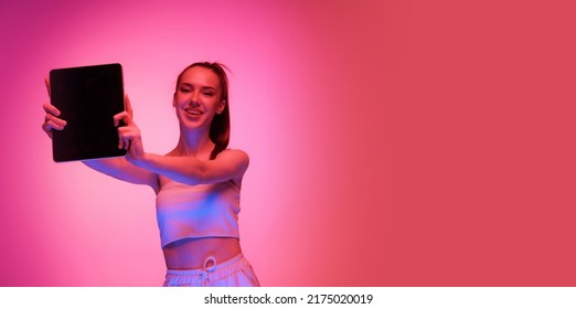 Showing Device Screen. One Young Charming Girl Using Digital Tablet Isolated On Magenta Color Background In Neon Light. Concept Of Beauty, Art, Fashion, Emotions, Youth. Copy Space For Ad