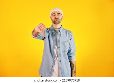 Showing Dislike. Studio Shot Of Smiliing Handsome Man In Jeans And White Hat On Yellow Background. Tatoo And Beard. 