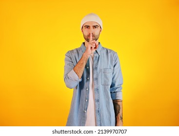 Showing Hush Sing. Studio Shot Of Smiliing Handsome Man In Jeans And White Hat On Yellow Background. Tatoo And Beard. 