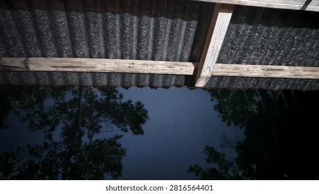 showing an inverted view showing the reflection of trees and the sky in a dark, still body of water, beneath a corrugated metal roof and wooden support beams.