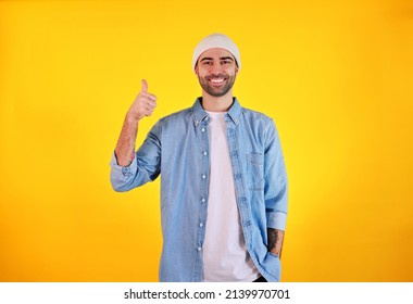 Showing Like. Studio Shot Of Smiliing Handsome Man In Jeans And White Hat On Yellow Background. Tatoo And Beard. Happy Concept.