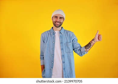 Showing Like. Studio Shot Of Smiliing Handsome Man In Jeans And White Hat On Yellow Background. Tatoo And Beard. Happy Concept.