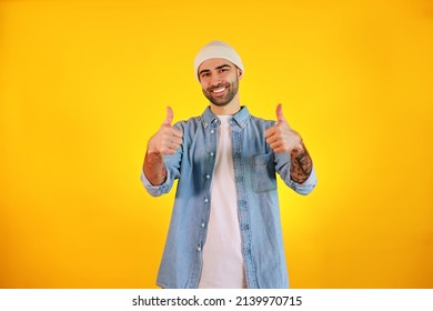 Showing Like. Studio Shot Of Smiliing Handsome Man In Jeans And White Hat On Yellow Background. Tatoo And Beard. Happy Concept.