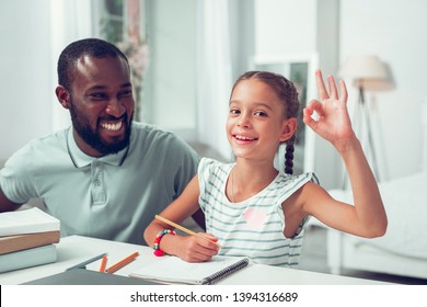 Showing ok sign. Happy cheerful cute sweet dark-haired Afro-American girl showing the ok sign while her handsome dark-haired bearded beaming smiling dad sitting near.