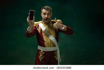 Showing phone with blank screen. Young man in suit as Nicholas II on dark green background. Retro style, comparison of eras concept. Beautiful male model like historical character, monarch, old
