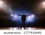 Showman. Middle Age Male entertainer, presenter or actor on stage. Arms to sides, smoke on background of spotlight. Rear view portrait of a male public speaker speaking at the microphone, pointing