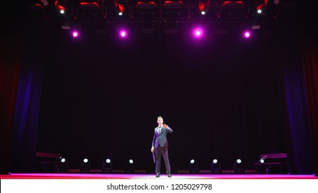 Showman. Young Male Entertainer, Presenter Or Actor On Stage. Back, Arms To Sides, Smoke On Background Of Spotlight. Rear View Of A Male Public Speaker Speaking At The Microphone, Pointing, In The.