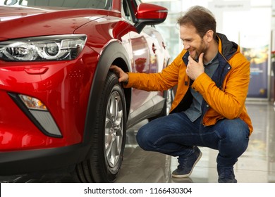 Showroom Of Modern Car Dealership. Male Customer Of Car Center Holding Hand On Wheel Of Automobile. Handsome Man Thinking About Characteristics Of New Red Car He Want Buy.   