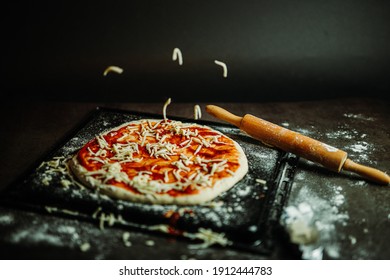 Shredded Mozzarella Cheese Falling On Home Made Pizza Dough With Tomato Sauce. Black Background, Wooden Rolling Pin And Flour On Setting. Messy Pizza Baking.