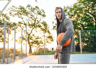 Shrewd Sunlit European Young Sporty Sportsman Man 20s Wear Grey Sports Clothes Hood Look Camera Training Hold In Hand Ball Play At Basketball Game Playground Court. Outdoor Courtyard Sport Concept.