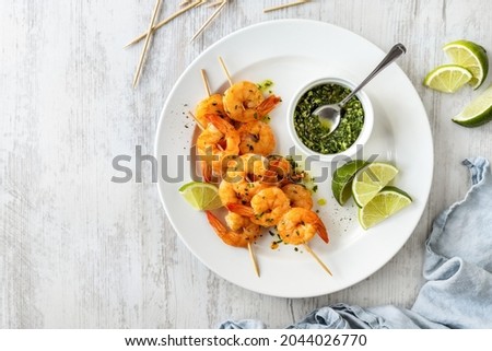 Shrimp skewers grilled, served  with green homemade pesto and lime slices on white plate top view on wooden table 