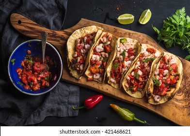 Shrimp tacos with homemade salsa, limes and parsley on wooden board over dark background. Top view. Mexican cuisine