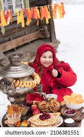 Shrovetide Concept Maslenitsa . Joyful Little Girl Bites A Large Bagel, Sits At A Table With A Samovar And Pancakes. Outdoor In Winter Time.