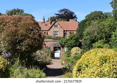 Shrubs And Trees Either Side Of A Gravel Path Lead Up To An Open Gateway And Onwards To A Large House. The Sky Is Blue And Cloudless