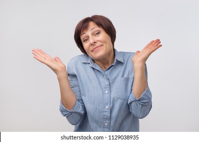 Shrugging Mature Woman In Doubt Looking At Camera. Attractive Confused Female Gesturing Do Not Know Sign On Gray Background