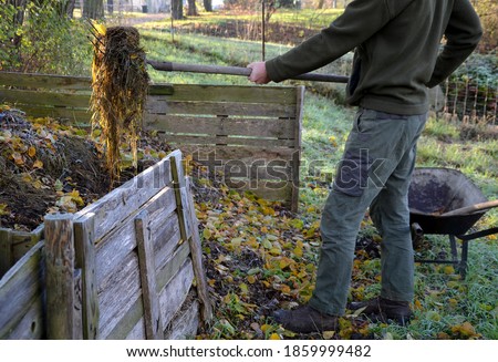 shuffling compost for aeration and better rot. the gardener uses a shovel and pitchfork in his hand. He has green pants and a jacket. Compost is in a wooden plank box. garden wheelbarrow