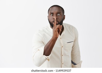 Shut Up And Listen. Pissed Angry African American Guy With Beard In White Shirt, Bending Towards Camera With Scary Expression, Saying Shh While Showing Shush Gesture With Index Finger Over Mouth