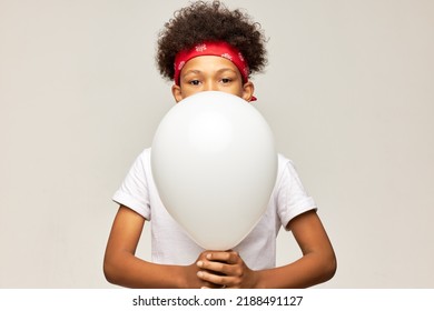 Shy African American Kid With Afro Hair And Red Bandana On Head Covering Face With Or Hiding Behind White Mockup Helium Balloon With Copy Space For Your Promotional Tex Or Content On Gray Background