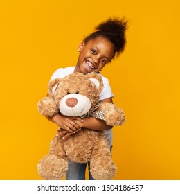 Shy African American Little Girl Embracing Her Teddy Bear And Smiling, Orange Studio Background